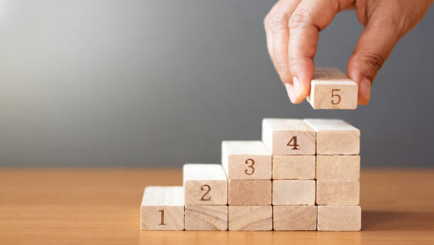 Women hand putting a wooden block on top and arranging wooden blocks stacking on wooden table in the shape of a staircase, Business concept for growth success process.